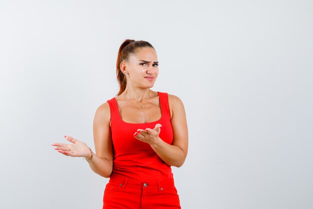 Giovane donna scontenta di una domanda stupida in canotta rossa, pantaloni e sguardo perplesso, vista frontale.