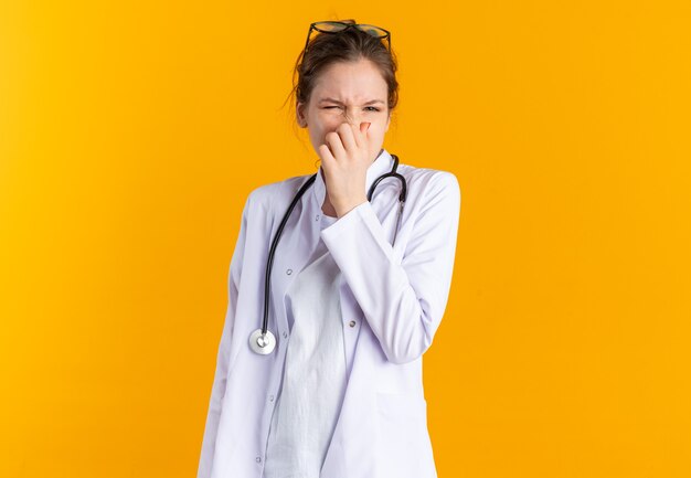 Giovane donna scontenta in uniforme da medico con uno stetoscopio che le chiude il naso