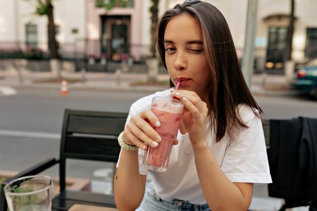 Giovane donna seducente che strizza l'occhio alla telecamera e sorride mentre beve frullato estivo e si siede sulla terrazza in primo piano Le curve nude e sottili delle clavicole sono sorprendenti