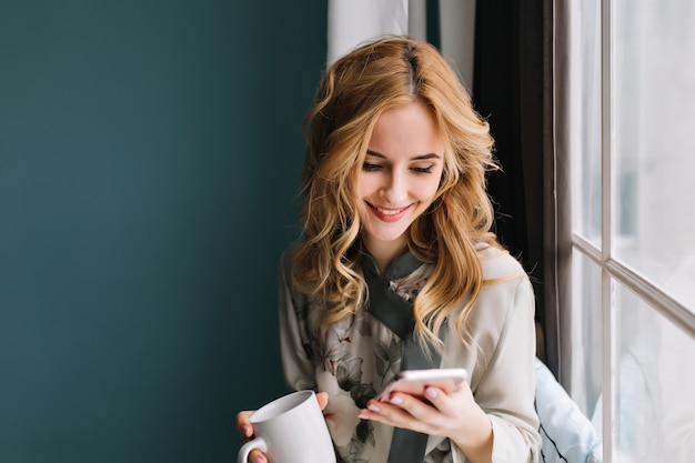 Giovane donna seduta accanto alla finestra e scrivere messaggi sul telefono mentre beve il caffè o il tè del mattino. I suoi capelli biondi sono ondulati. È in una stanza luminosa con pareti turchesi. Indossare un pigiama di seta.