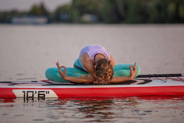 Giovane donna seduta sul paddle board, praticando la posa di yoga. Fare esercizi di yoga a bordo sup, riposo estivo attivo. Esercizio per la flessibilità e l'allungamento dei muscoli.