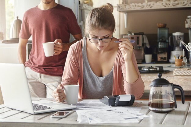 Giovane donna seria e concentrata con gli occhiali che tiene la tazza in una mano e la penna nell'altra, concentrata sul lavoro di ufficio