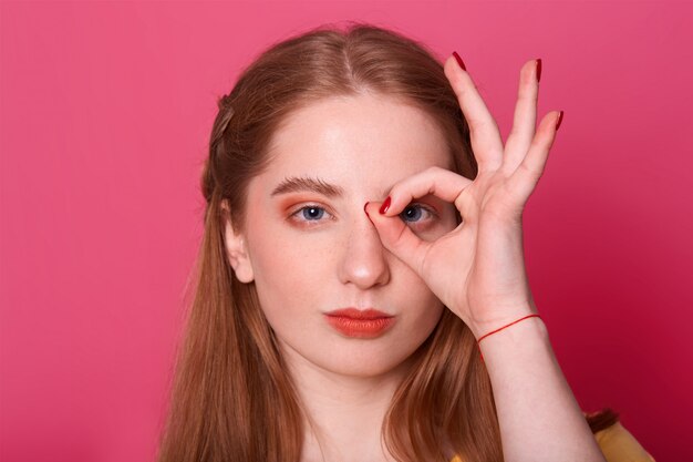 giovane donna seria, fa segno giusto, contro i suoi occhi, esprime sicurezza, modella posa sul rosa, fotografa. Concetto di persone e gesti.