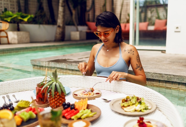 Giovane donna sexy con il tatuaggio in costume da bagno facendo colazione in una piscina privata. Ragazza rilassante in piscina bevendo caffè e mangiando frutta. Piatto di frutta, frullato a bordo piscina.