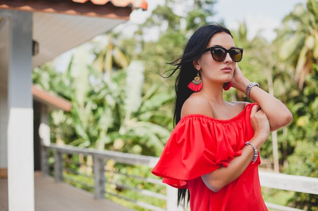 Giovane donna sexy elegante in abito estivo rosso in piedi sulla terrazza in hotel tropicale, sfondo di palme, capelli neri lunghi, occhiali da sole, orecchini etnici, occhiali da sole, sorridente