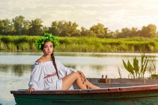 Giovane donna sexy in barca al tramonto. La ragazza ha una corona di fiori in testa, si rilassa e naviga sul fiume. Bel corpo e viso. Fotografia artistica di fantasia. Concetto di bellezza femminile, riposa nel male