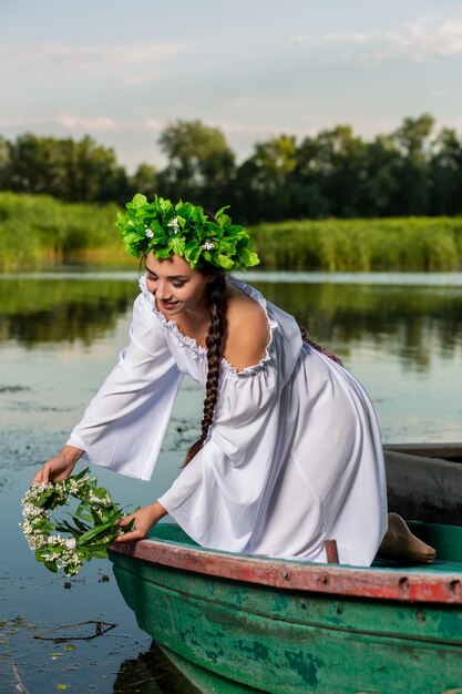 Giovane donna sexy in barca al tramonto. La ragazza ha una corona di fiori in testa, si rilassa e naviga sul fiume. Bel corpo e viso. Fotografia artistica di fantasia. Concetto di bellezza femminile, riposa nel male