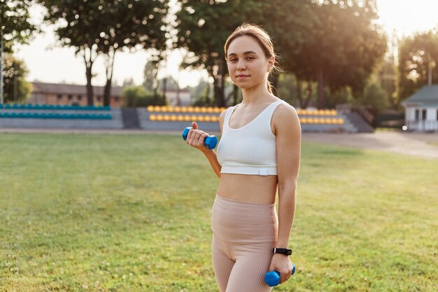 Giovane donna sicura di sé adulta che indossa top bianco sportivo e leggins beige in piedi nello stadio con manubri in mano, guardando direttamente la telecamera, allenarsi all'aperto.