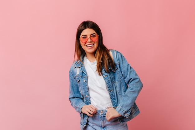 Giovane donna sicura in jeans che sorride alla macchina fotografica. Vista frontale della ragazza bruna in occhiali da sole isolato su sfondo rosa.