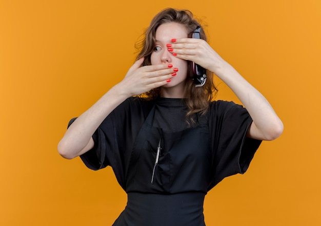 Giovane donna slava barbiere che indossa l'uniforme guardando il lato tenendo i tagliacapelli isolati su sfondo arancione