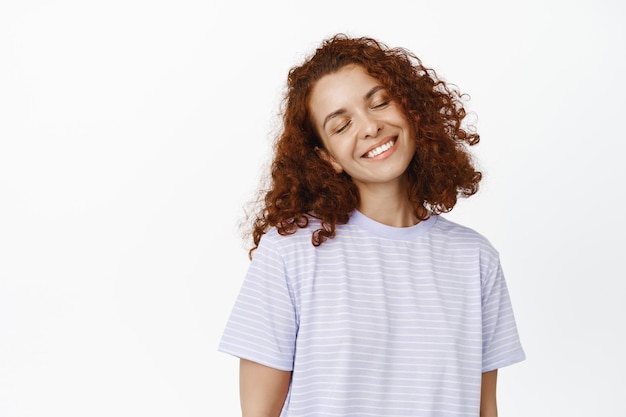 Giovane donna sognante con i capelli ricci rossi, l'imaging smth, la fantasia, gli occhi clsoe e sorridente felice, in piedi in t-shirt su sfondo bianco.