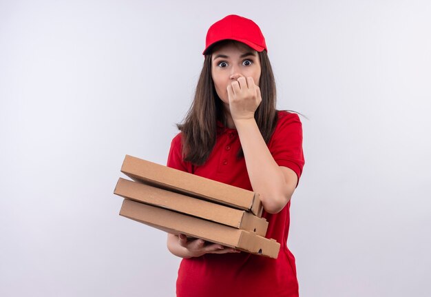 Giovane donna sorpresa di consegna che indossa la maglietta rossa in berretto rosso che tiene una scatola di pizza sul muro bianco isolato