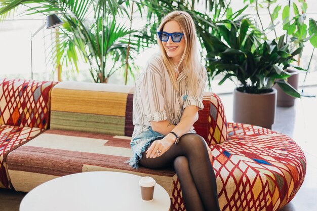 Giovane donna sorridente abbastanza elegante che si siede nel caffè hipster sul divano che indossa camicia e occhiali da sole blu