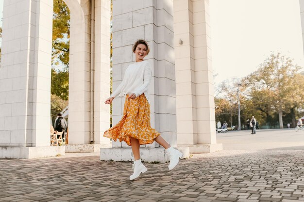 Giovane donna sorridente abbastanza felice in vestito stampato giallo e maglione bianco lavorato a maglia