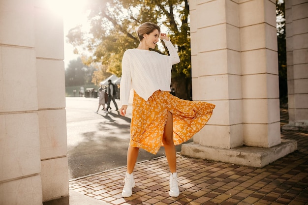 Giovane donna sorridente abbastanza felice in vestito stampato giallo e maglione bianco lavorato a maglia