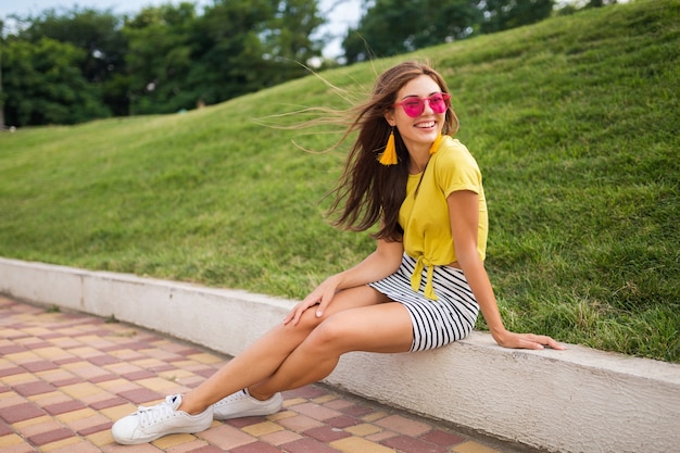 Giovane donna sorridente alla moda attraente che si diverte nel parco cittadino, positivo, emotivo, indossa un top giallo, minigonna a righe, occhiali da sole rosa, scarpe da ginnastica bianche, tendenza della moda in stile estivo, gambe lunghe