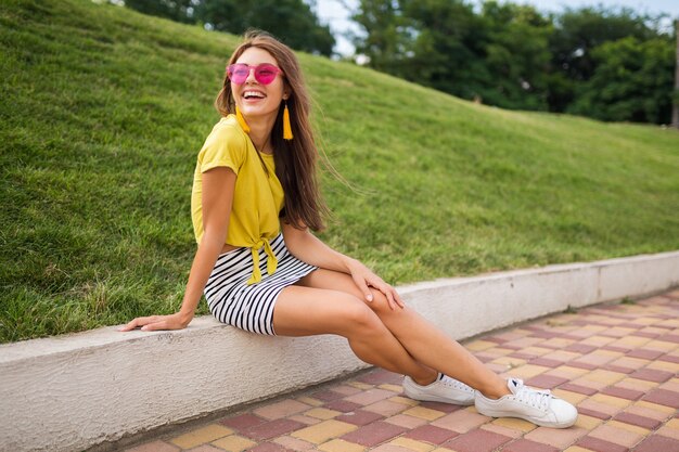 Giovane donna sorridente alla moda attraente che si diverte nel parco cittadino, positivo, emotivo, indossa un top giallo, minigonna a righe, occhiali da sole rosa, scarpe da ginnastica bianche, tendenza della moda in stile estivo, gambe lunghe