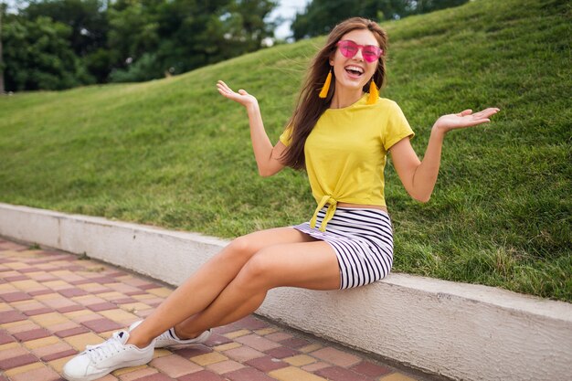 Giovane donna sorridente alla moda che si diverte nel parco cittadino, positiva, emotiva, indossa un top giallo, minigonna a righe, occhiali da sole rosa, scarpe da ginnastica bianche, tendenza della moda in stile estivo, felice, alzando le mani