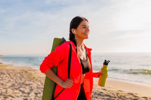 Giovane donna sorridente attraente che fa sport all'alba di mattina sulla spiaggia del mare che tiene stuoia di yoga e bottiglia di acqua, stile di vita sano, ascolto di musica sugli auricolari, giacca a vento rosa da indossare