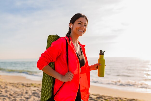 Giovane donna sorridente attraente che fa sport all'alba di mattina sulla spiaggia del mare che tiene stuoia di yoga e bottiglia di acqua, stile di vita sano, ascolto di musica sugli auricolari, giacca a vento rosa da indossare