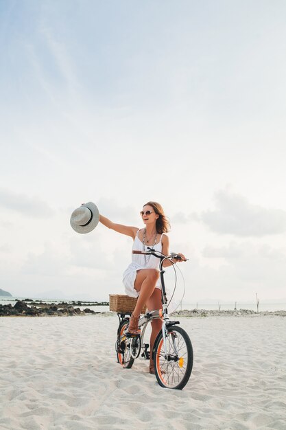 Giovane donna sorridente attraente in vestito bianco che guida sulla spiaggia tropicale in bicicletta che indossa cappello e occhiali da sole