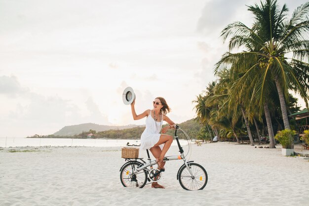 Giovane donna sorridente attraente in vestito bianco che guida sulla spiaggia tropicale in bicicletta che indossa cappello e occhiali da sole