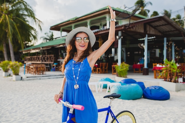 Giovane donna sorridente attraente in vestito blu che cammina sulla spiaggia tropicale con la bicicletta che indossa cappello e occhiali da sole