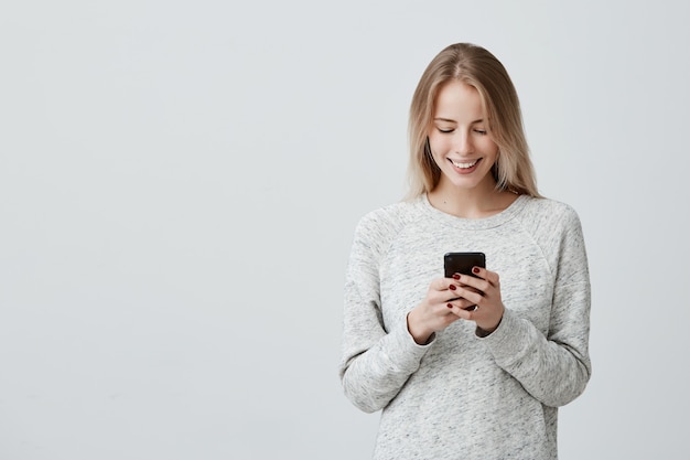 Giovane donna sorridente bionda che dimostra i denti bianchi facendo uso del telefono cellulare, messaggio