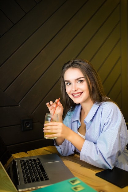 Giovane donna sorridente che beve bevanda fredda nella caffetteria con il computer portatile