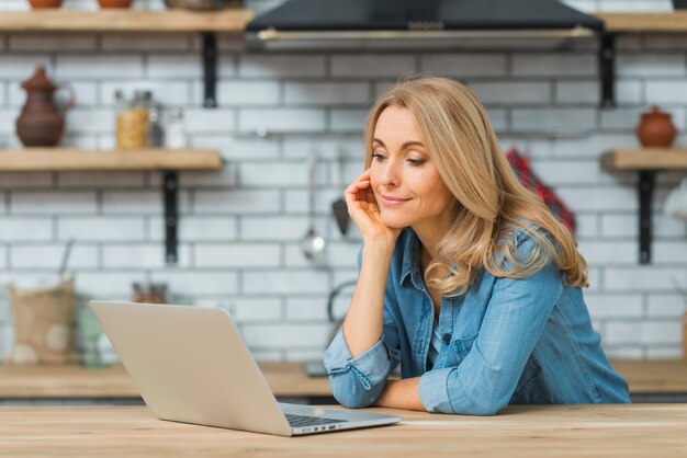 Giovane donna sorridente che esamina computer portatile sulla tavola nella cucina