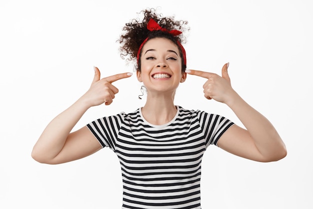 Giovane donna sorridente che mostra i suoi denti bianchi perfetti, indicando la bocca e guardando felice, in piedi in t-shirt su sfondo bianco