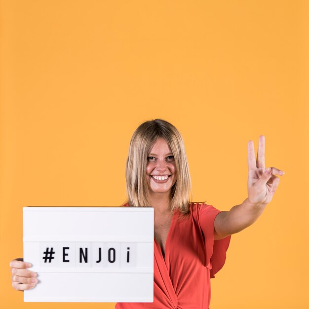 Giovane donna sorridente che mostra il segno di pace mentre tenendo scatola leggera con testo