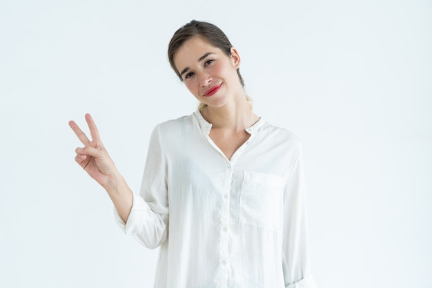 Giovane donna sorridente che mostra il segno di vittoria, inclinando testa a spalla e rivolto verso l&#39;obiettivo.