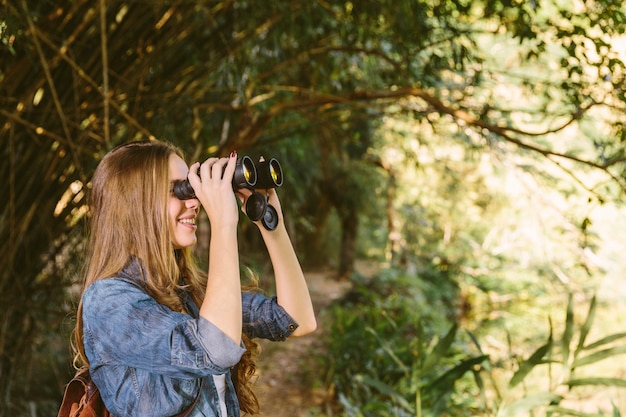 Giovane donna sorridente che osserva tramite il binocolo nella foresta