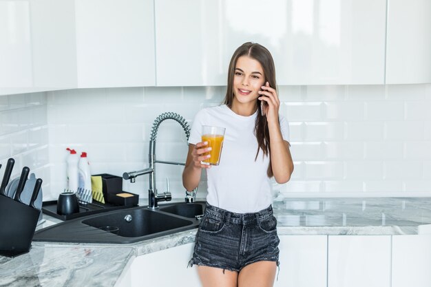 Giovane donna sorridente che parla sul telefono cellulare mentre stando su una cucina con bicchiere di succo