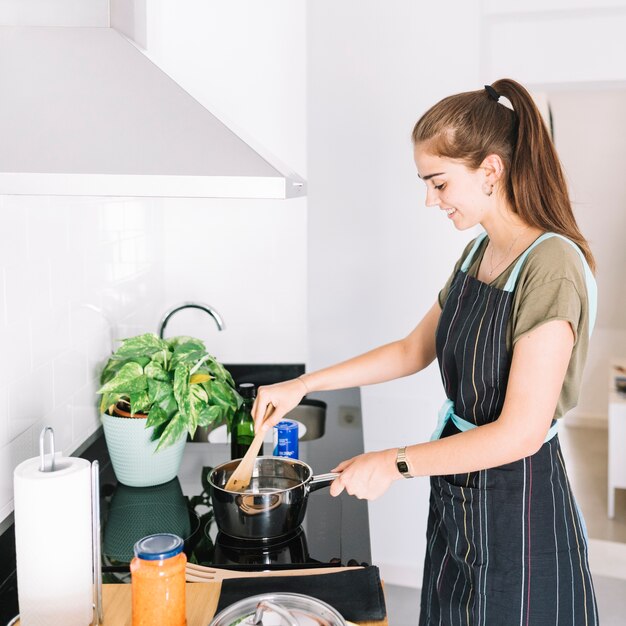 Giovane donna sorridente che prepara alimento nella casseruola sulla stufa elettrica