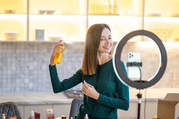 Giovane donna sorridente che presenta la lacca per capelli durante il tutorial di bellezza online