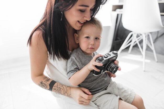 Giovane donna sorridente che si siede con la sua macchina fotografica della tenuta del figlio a casa