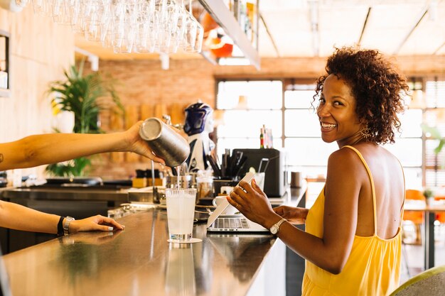 Giovane donna sorridente che si siede vicino al contatore della barra e barista che prepara cocktail