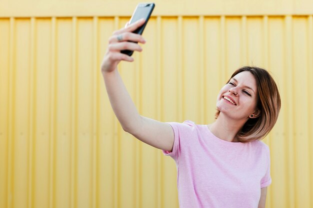 Giovane donna sorridente che sta contro la lamina di metallo gialla ondulata che prende selfie sul telefono cellulare