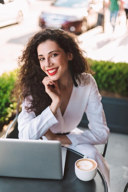Giovane donna sorridente con i capelli ricci scuri in costume bianco seduta al tavolo con una tazza di caffè e laptop mentre guarda felicemente in camera nella caffetteria sulla strada