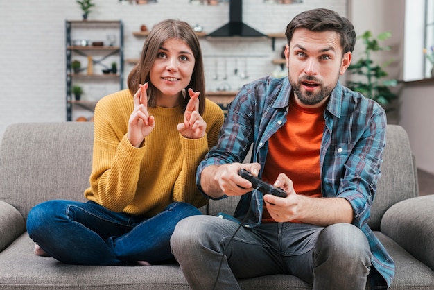 Giovane donna sorridente con le dita attraversate che si siedono vicino all&#39;uomo che gioca video gioco