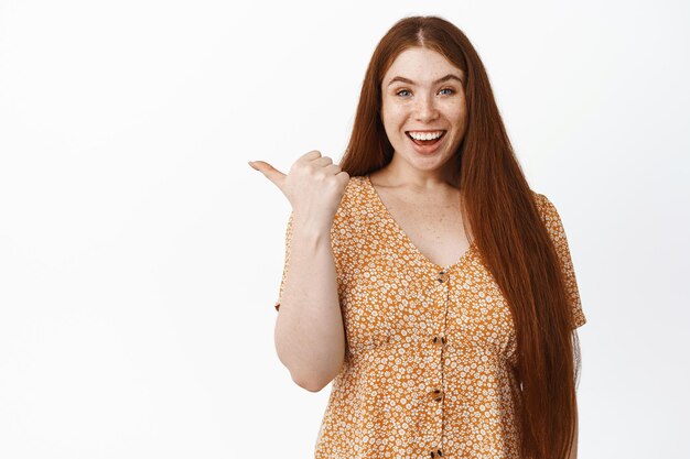 Giovane donna sorridente con lunghi capelli rossi che punta il dito a sinistra e che sembra entusiasta che mostra la pubblicità in piedi su sfondo bianco