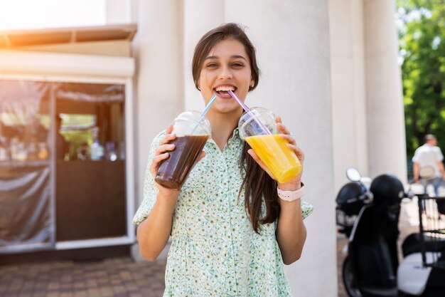 Giovane donna sorridente e beve due cocktail con ghiaccio in bicchieri di plastica con paglia sulla strada cittadina.