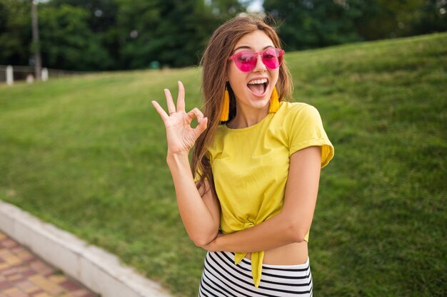 Giovane donna sorridente elegante attraente che si diverte nel parco cittadino, positivo, emotivo, indossa un top giallo, minigonna a righe, occhiali da sole rosa, tendenza della moda in stile estivo, mostrando segno ok, colorato