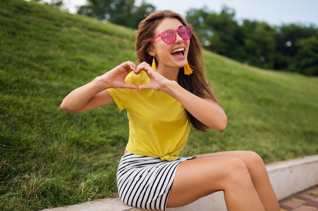 Giovane donna sorridente felice abbastanza elegante che si diverte nel parco cittadino, positiva, emotiva, indossa un top giallo, minigonna a righe, occhiali da sole rosa, tendenza della moda in stile estivo, mostrando il segno del cuore