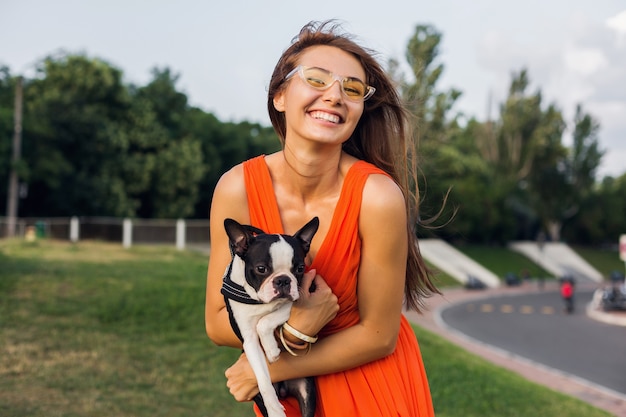 Giovane donna sorridente felice che tiene il cane di boston terrier nel parco, giornata di sole estivo, umore allegro, giocando con animali, abbracci, vestito arancione, occhiali da sole, stile estivo