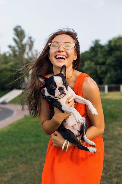 Giovane donna sorridente felice che tiene il cane di boston terrier nel parco, giornata di sole estivo, umore allegro, giocando con animali, abbracci, vestito arancione, occhiali da sole, stile estivo