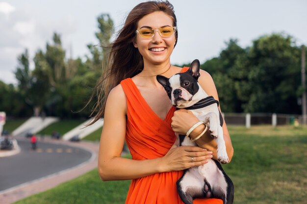 Giovane donna sorridente felice in vestito arancione divertendosi a giocare con il cane nel parco, stile estivo, umore allegro