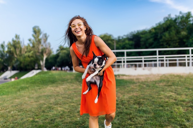 Giovane donna sorridente felice in vestito arancione divertendosi a giocare con il cane nel parco, stile estivo, umore allegro